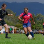 Women soccer players in action during a match on a lush green field with a mountain backdrop.