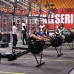 Competitors row on machines during an indoor fitness event. Focus on teamwork and endurance.