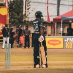 A cricket player stands ready on the field during a sunny match, surrounded by spectators.