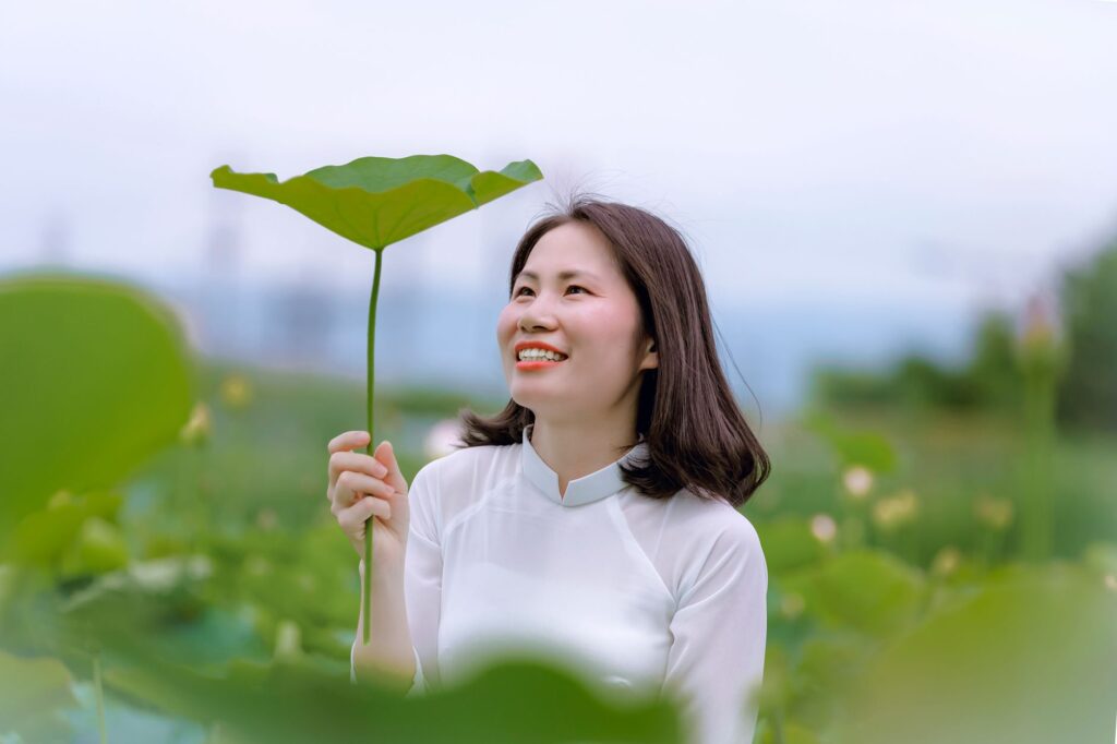 Smiling woman holding a lotus leaf, enjoying a sunny outdoor setting.