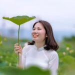 Smiling woman holding a lotus leaf, enjoying a sunny outdoor setting.