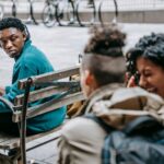 Back view of young friends in casual outfit with backpacks laughing and gossiping behind back of African American male sitting on bench in headphones in city street in daylight