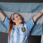 Joyful woman holding Argentina flag wearing national jersey indoors.