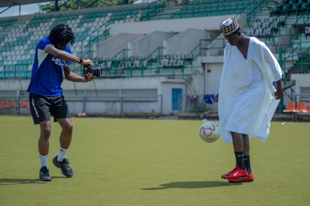 Two young men enjoy a casual soccer game in traditional attire at an Abuja stadium.