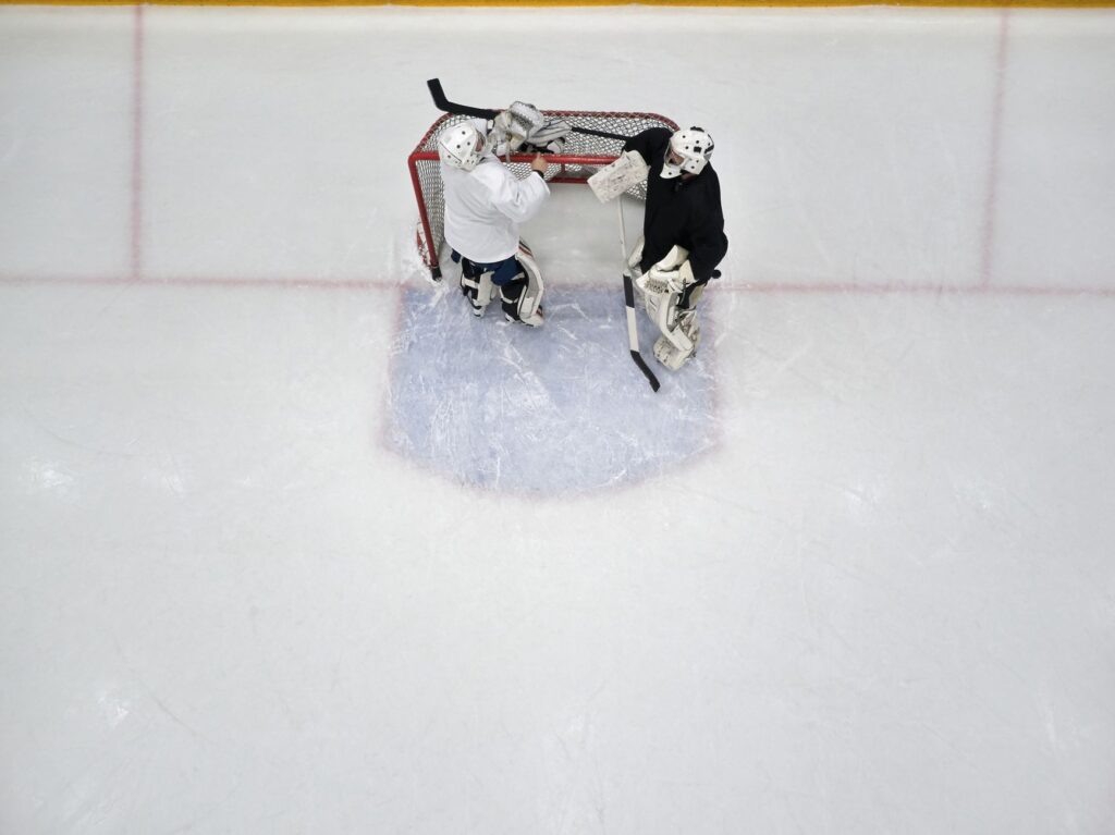 Top view of two ice hockey goalies in gear, standing in front of the net on an indoor rink during practice.