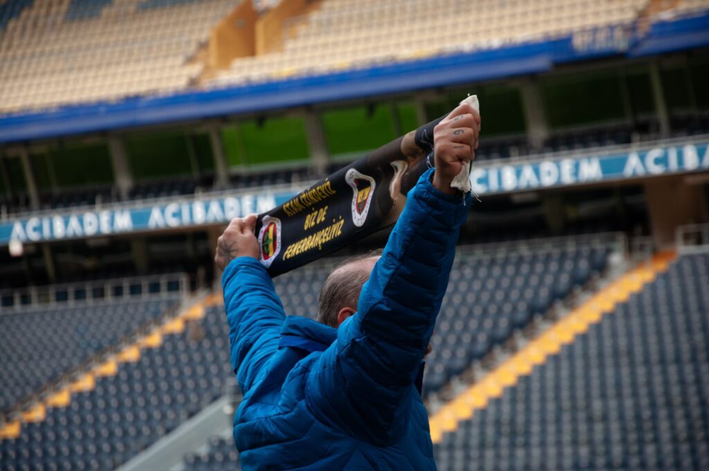 A lone fan in blue jacket raises scarf in empty stadium supporting team.
