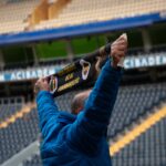 A lone fan in blue jacket raises scarf in empty stadium supporting team.