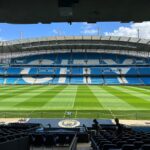 Panoramic view of Etihad Stadium, showcasing the empty football pitch and stands under a clear blue sky.