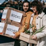 Joyful couple carrying boxes and plants while relocating to a new neighborhood on a sunny day.
