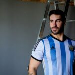 An adult male wearing an Argentina soccer jersey stands beside a ladder indoors.