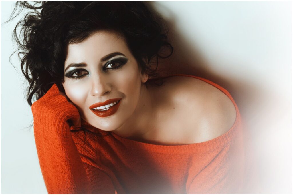 Woman with bold makeup and messy hair posing in a red sweater.