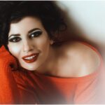 Woman with bold makeup and messy hair posing in a red sweater.