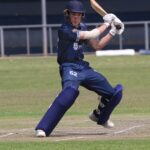 Batsman in blue uniform playing a shot on a grassy cricket field during a sunny day.