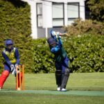 Action shot of a cricket batsman playing a shot while the wicketkeeper looks on during a match outdoors.