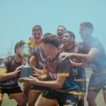 A joyful sports team celebrating their victory with a trophy outdoors on a sunny day.