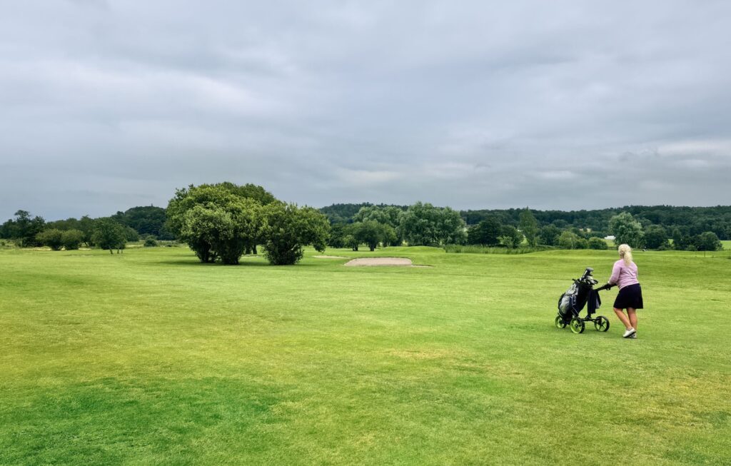 A woman strolls across Solängen golf course in Sweden, pushing a golf cart on a cloudy day.