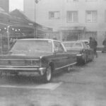 Black and white photo of classic cars parked in a city market setting, evoking a vintage feel.