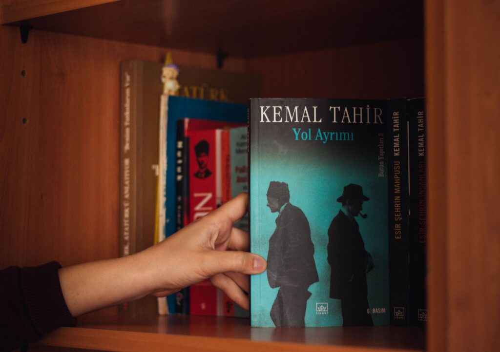 Crop unrecognizable female choosing book with illustration of men in black wear on wooden bookcase near multicolored collection of literature in apartment