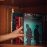 Crop unrecognizable female choosing book with illustration of men in black wear on wooden bookcase near multicolored collection of literature in apartment