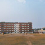 Daytime cricket match on a large sports field in Delhi, India with players and floodlights.