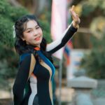 Smiling woman in colorful dress posing with hand raised in a sunny outdoor setting.