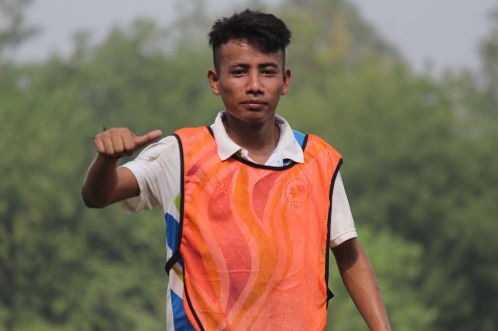 A young male athlete in an orange sports vest gives a thumbs-up in a green outdoor setting.