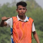 A young male athlete in an orange sports vest gives a thumbs-up in a green outdoor setting.