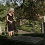 A senior woman standing in a cemetery, holding flowers and expressing grief.
