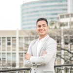 Confident man in a light suit smiling in a city environment with modern buildings.