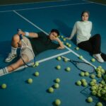Young adults sit with tennis rackets and balls on an outdoor court, enjoying a relaxed moment.