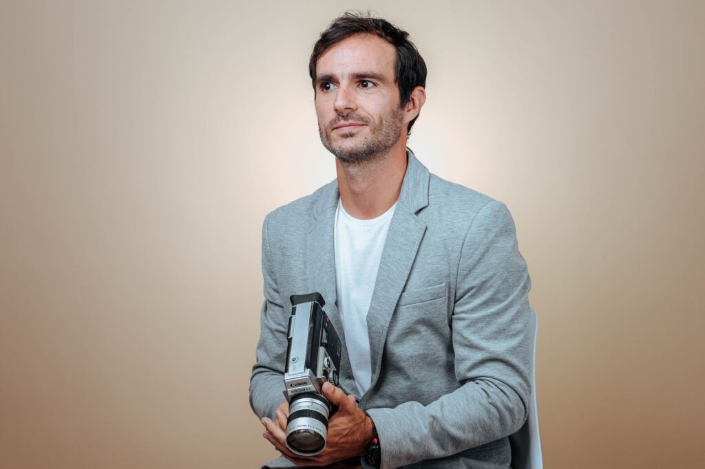 Man sitting with a vintage camera against a beige background in studio lighting.