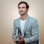 Man sitting with a vintage camera against a beige background in studio lighting.