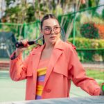 Fashionable young woman in a vibrant coat posing confidently on a tennis court.