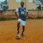 A teenage soccer player in uniform poses with a ball on a dirt field.