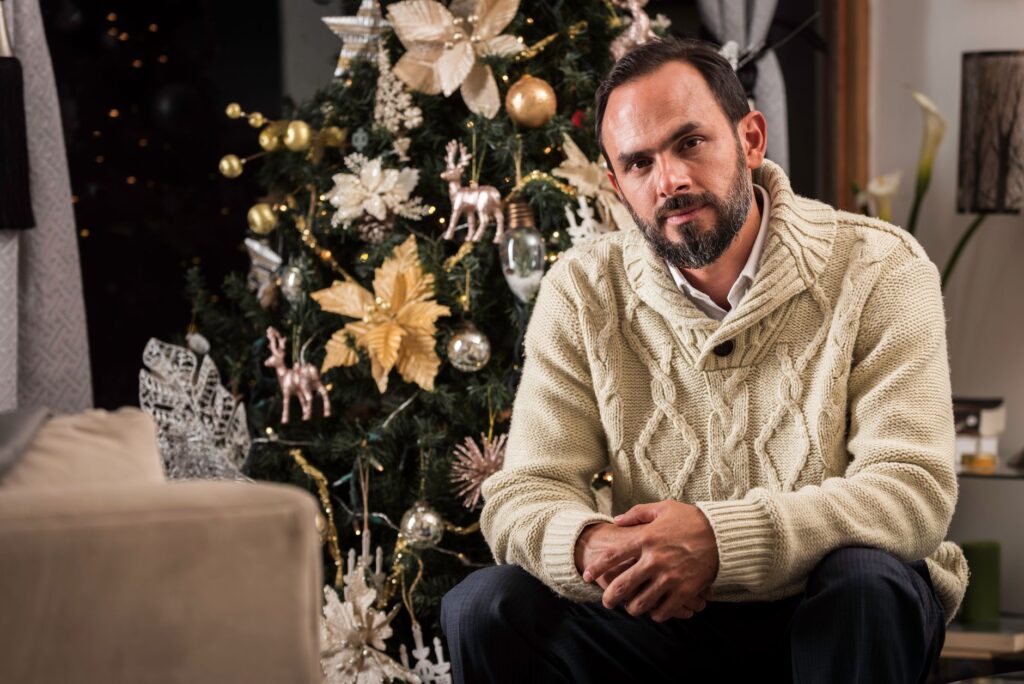 Man in cozy sweater posing by a decorated Christmas tree indoors, creating a festive atmosphere.
