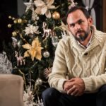 Man in cozy sweater posing by a decorated Christmas tree indoors, creating a festive atmosphere.