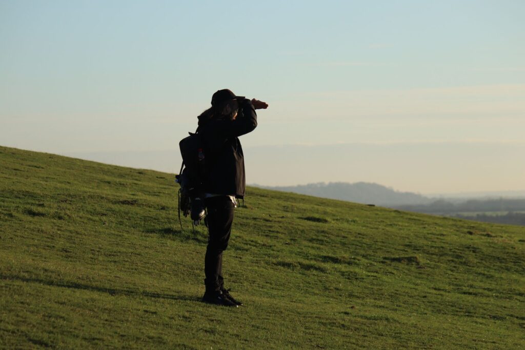 A person stands on a grassy hill, looking towards the horizon on a clear day.