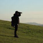 A person stands on a grassy hill, looking towards the horizon on a clear day.