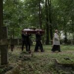 A solemn funeral procession with a priest and pallbearers carrying a coffin through a lush cemetery.