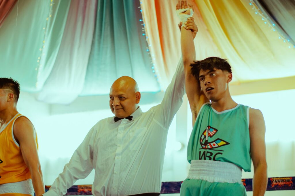 A triumphant teenage boxer winning a match, his hand raised by a referee, in a vibrant indoor setting.