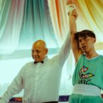A triumphant teenage boxer winning a match, his hand raised by a referee, in a vibrant indoor setting.
