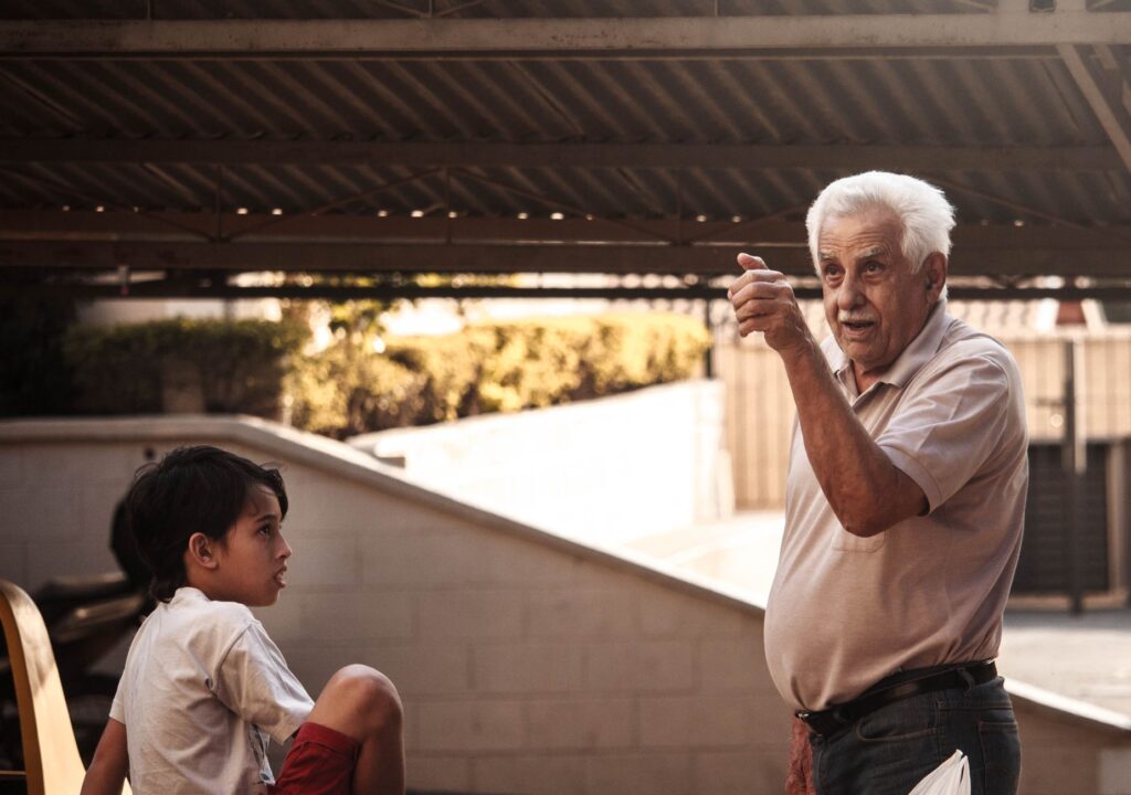 A senior adult gesturing and talking to a young boy outdoors under a canopy.