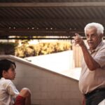 A senior adult gesturing and talking to a young boy outdoors under a canopy.