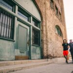 A police officer escorts a handcuffed individual past a brick building.