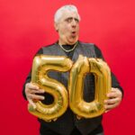 A joyful senior holding gold '50' balloons against a vibrant red backdrop.