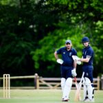 Two cricket players in gear discussing strategy on a sunny day outdoors, ready to play.