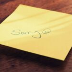 A yellow sticky note with a handwritten apology message and smiley face on a wooden surface.