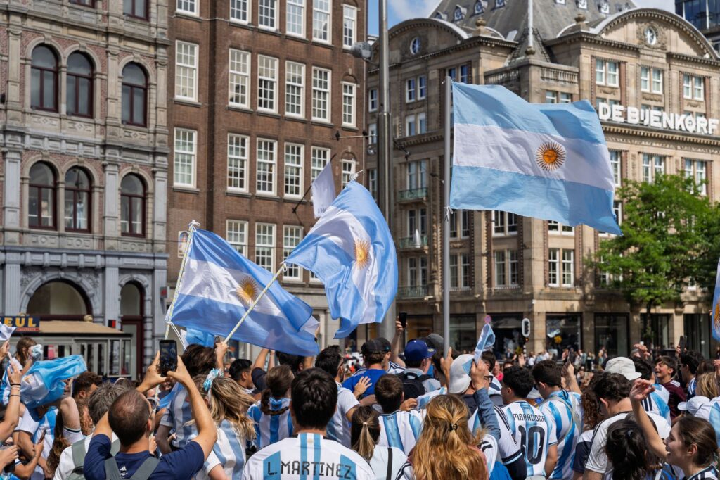 Lively crowd of Argentinian soccer fans waving flags in a bustling city square celebrating victory.