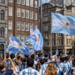 Lively crowd of Argentinian soccer fans waving flags in a bustling city square celebrating victory.
