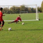 A soccer player prepares to take a shot as the goalkeeper dives to save in an outdoor match.